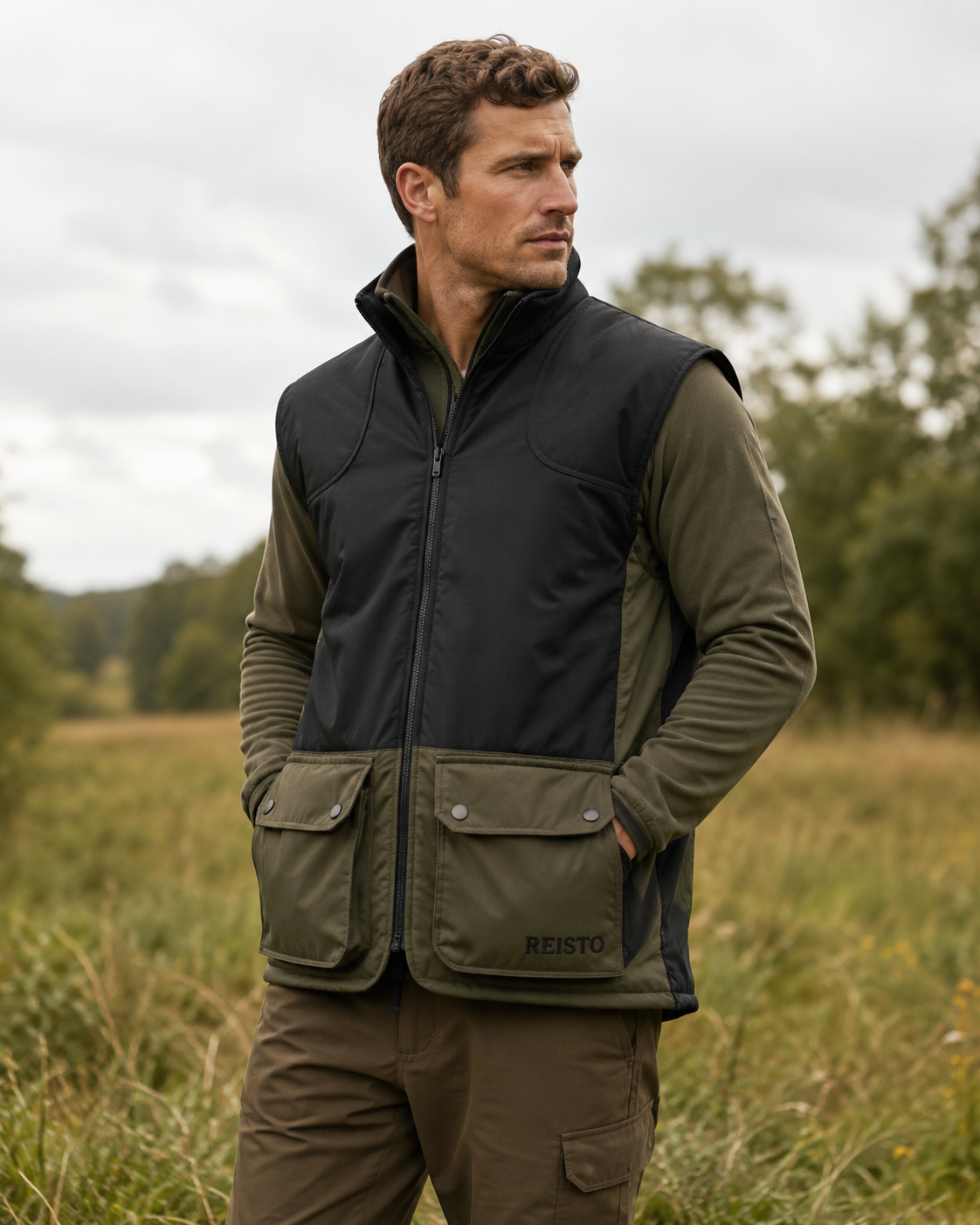 A man wearing a Reisto outdoor vest over an olive green long-sleeve shirt, standing in a grassy field under an overcast sky.